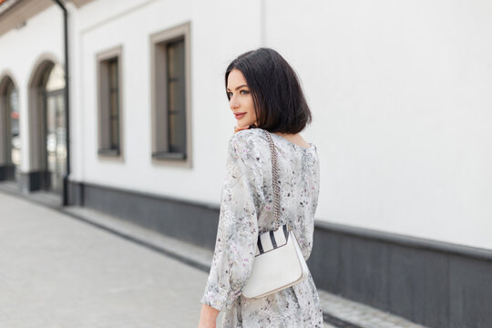 Pretty fashion summer woman with hairstyle in vintage stylish flowers dress with white bag walks in the city near a white building. Urban spring female style look