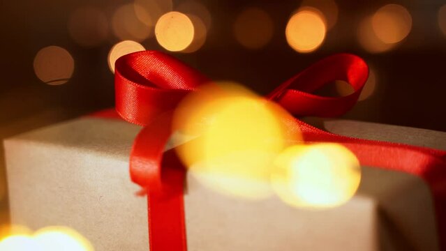 Close up Christmas gift craft paper box with red ribbon bow over dark lights bokeh background. New year eve holiday composition with present handmade box. Eve preparation. Family time, cozy home