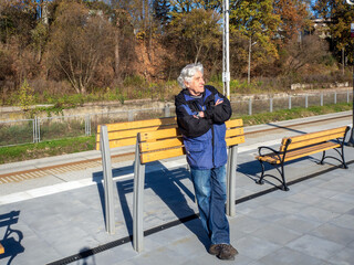 Elderly gray-haired senior in blue jeans leaning against a wooden modern leaning bench or leaning stand or lean bar (no established name yet) waiting for the train on a small railway station in Poland - 543641980