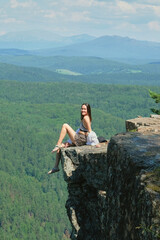 A girl with long hair sits on the edge of a cliff. Landscape, valley, mountains, summer, forest, river. Panorama, height.