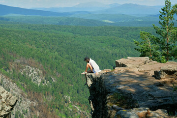 A girl with long hair sits on the edge of a cliff. Landscape, valley, mountains, summer, forest, river. Panorama, height.