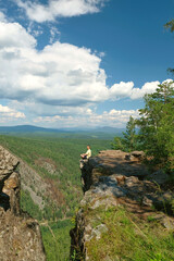 A man in a yellow T-shirt sits on the edge of a cliff. Landscape, valley, mountains, summer, forest, river. Panorama, height.