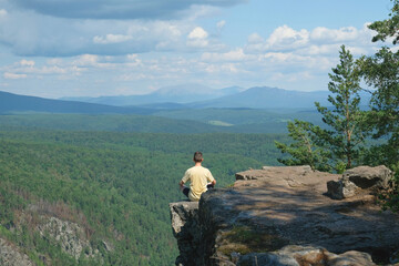 A man in a yellow T-shirt sits on the edge of a cliff. View from the back. Meditation. Landscape, valley, mountains, summer, forest, river. Panorama, height.