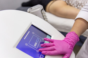 A medic adjusts the operating mode of the anti-cellulite massage machine with compression and vibration technology in beautician office.
