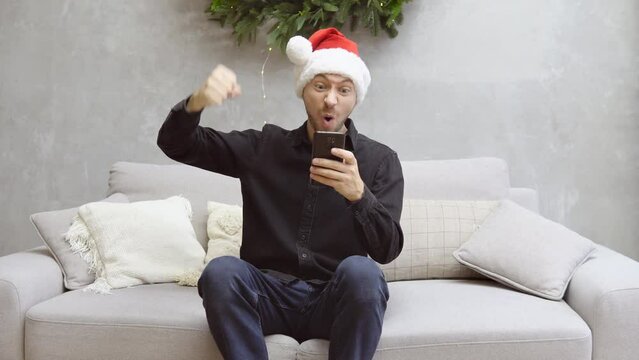 Portrait of young emotional man wearing christmas santa hat using smartphone very happy and rejoices