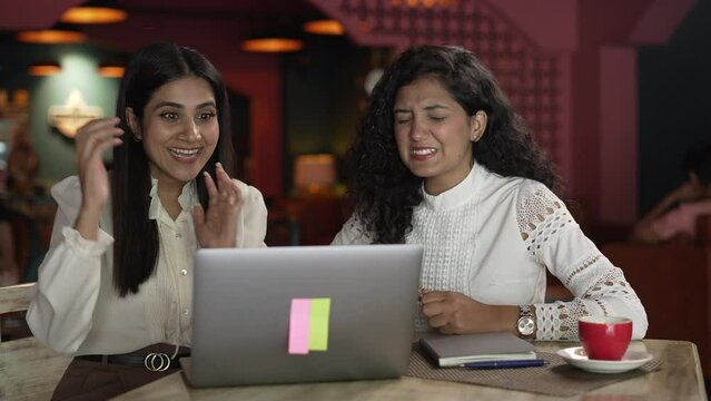 Attractive two young Asian women friends supporting different team on laptop.Indian female siting on upset with loser football game scored and other girl wining at the coffee shop.
