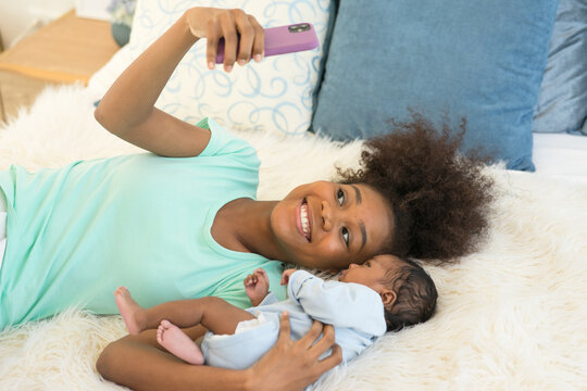 Young mother African American and newborn baby making video call and smiling greet. Happy elder sister taking Selfie with little brother infant lying on bed looking at mobile phone smiling carefree.