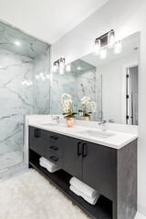 A beautiful bathroom with a dark vanity and white granite counter top. A plant and towels sit on the vanity. The shower is lined with marble tiles. Lights on.