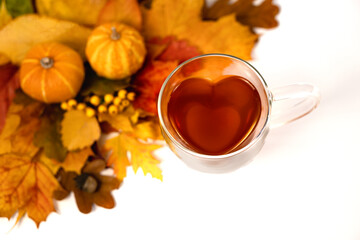 Herzförmige Tasse aus Glas mit Tee gefüllt auf weißem Hintergrund, Draufsicht, Laub, Eicheln und zwei kleine Kürbisse als Dekoration in der linken oberen Ecke, horizontal 