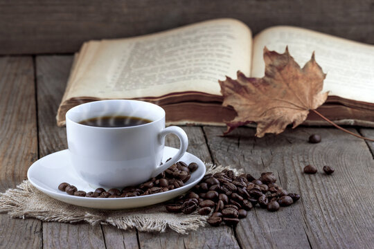 Close-up view of white cup of coffee with autumn leaf and old book on wooden background