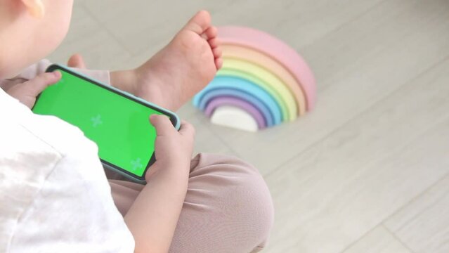 A small child sits on a chair next to a multicolored pyramid with bare legs and uses a mobile phone with a green screen.