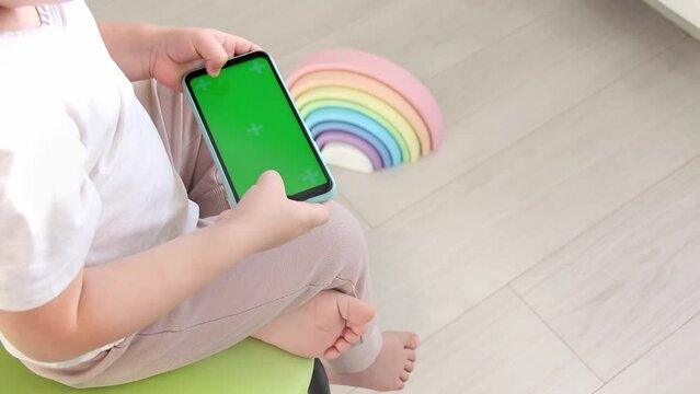 A small child sits on a chair next to a multicolored pyramid with bare legs and uses a mobile phone with a green screen.
