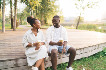Cheerful black couple having conversation while sitting on wooden walkway - obrazy, fototapety, plakaty