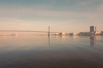 Fototapeta na wymiar Suspension Bridge and View of Riverfront on Opposite Shore on Hazy Day