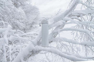 White thermo bottle for winter leisure. Flask thermos of drinks on snow covered tree on background of winter landscape