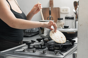 latina woman preparing a typical colombian breakfast, grabbing the arepa to turn it over while preparing the chocolate.