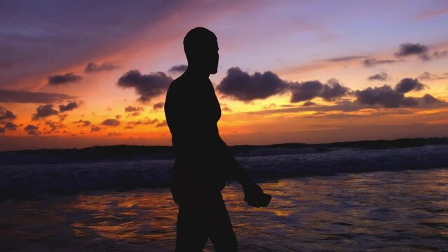 Silhouette of a man walking along the ocean against the backdrop of a bright fiery multi-colored sunset. Man enjoying the peace and sound of the waves. Slow motion 4k footage of a beautiful landscape