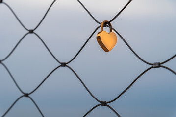 Padlock in the shape of a heart and gold color hooked on a metal fence as a sign of love for a couple.