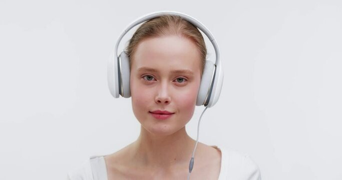 Portrait of young girl with headphones posing in silated white studio. Smiling gitl listening to music and looking at the camera, sitting in the white background