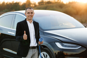 Fototapeta na wymiar Portrait of caucasian businessman standing near electric charging station looking at camera and showing thumb up. Male holds a charging cable type 2 mennekes in his hands. Eco-friendly transport