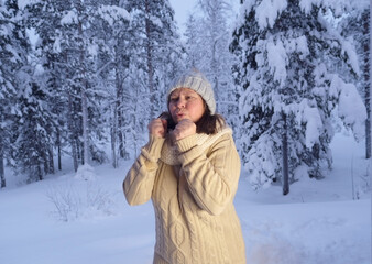Fototapeta na wymiar mature woman 50 years old with frozen hands try to warm them with breath in winter forest, early evening in park among snow-covered trees, beautiful landscape, concept active lifestyle, winter nature