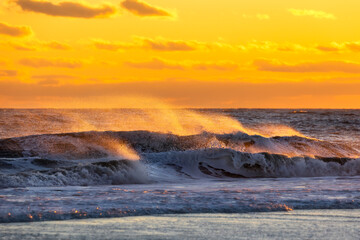 Spray from large waves in rough surf being lit up with golden light at sunset. Long Island New York