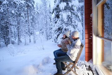 mature couple, man and woman are sitting on cozy wooden chairs on terrace of country house in winter forest and drinking hot tea, early evening, warm light from large window, enjoying winter nature