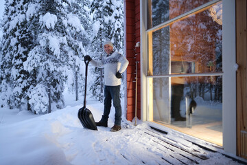 mature man in Scandinavian sweater cleans snow with snowshovel on terrace, snow-covered trees,...