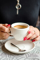 Woman in a black dress and red nails holding white cup of coffee over saucer in cafe
