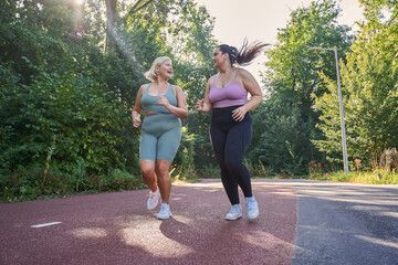 Positive overweight woman looking at her bestie and laughing while jogging at the street - obrazy, fototapety, plakaty