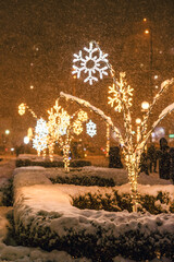 Snowy city park with shining and glowing snowflakes shape New year decorations at night. Festive and winter holidays season background.