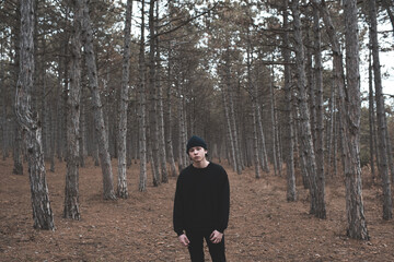 Teenage boy 16-17 year old walk in dark wooden wear black clothes and hat outdoor over nature...