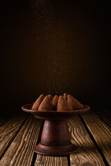 cocoa powder is poured from top on chocolate truffles in ceramic dishes against the background of a brown backdrop with a spot of light standing on a table of old boards. simple dark still life