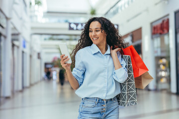 Happy and successful woman shopping for clothes in a supermarket store, Hispanic woman holding a smartphone reading online messages and browsing offers with discounts and sales