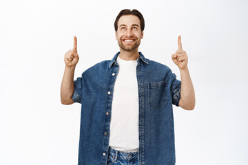 Handsome smiling man pointing fingers up, shows banner or advertisement on top, standing over white...
