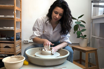 A female brunette potter in white shirt and blue jeans makes a bowl or vase on a fast spinning potter's wheel.
