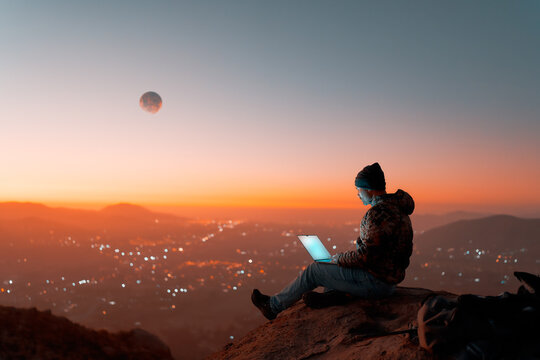 silhouette of digital nomad sitting on top of a hill working with his laptop over the city at dusk