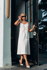 full length portrait of a fashionable old woman in white dress and sunglasses exits the door of the...