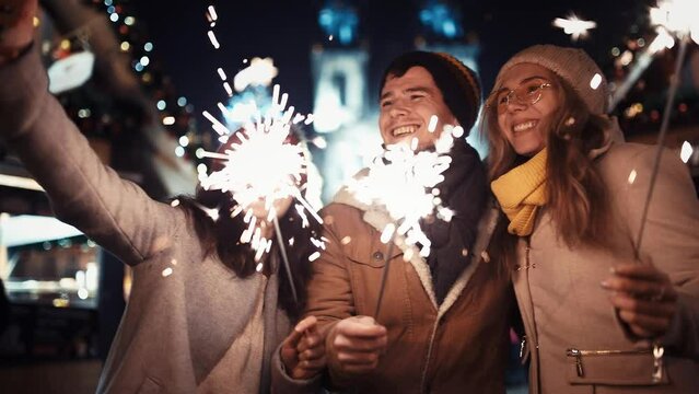 Three friends, a man and a girl with sparklers call on the phone on New Year's Eve by family video and cheerfully congratulate in the middle of the street in the lights of the Christmas market