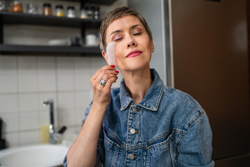 One woman mature caucasian female using gua sha rose quartz face massage stone in the kitchen at...