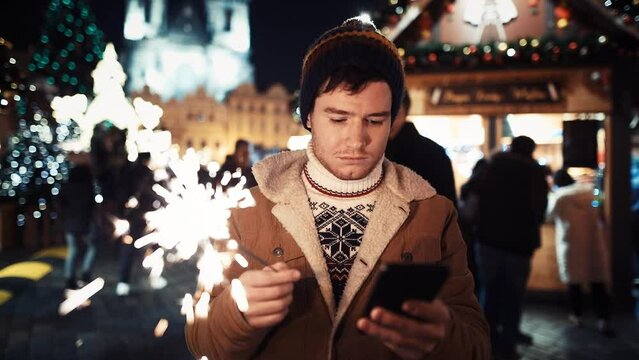 A sad young man with a dying sparkler is standing alone in the middle of a festive fair on New Year's Eve and trying to call someone from family or friends by phone. A lonely guy abandoned by a girl