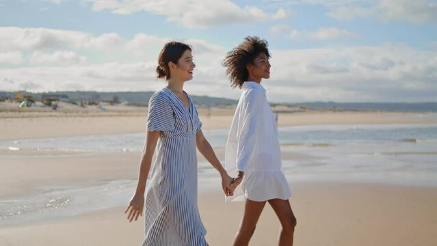 Joyful lesbian couple walking sea shore on sunny day. Happy girls holding hands