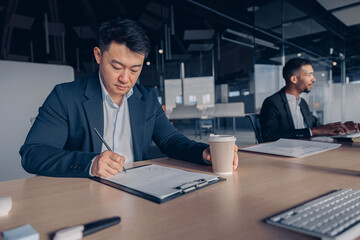 Focused asian businessman drinking coffee while working with documents in modern office