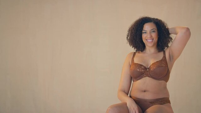 Studio shot of confident and positive woman wearing underwear sitting on stool looking into camera and smiling - shot in slow motion