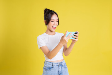 portrait of young beautiful asian girl holding phone playing game on yellow background