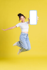 full body image of young Asian girl holding phone with cheerful face on yellow background