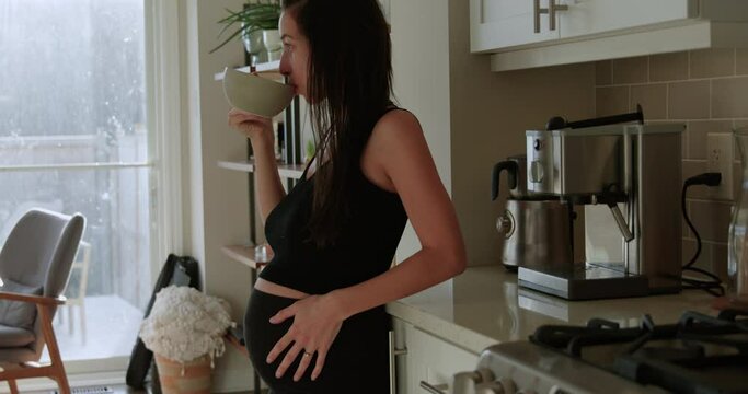 Pregnant woman second trimester drinks tea while leaning on kitchen counter - side profile