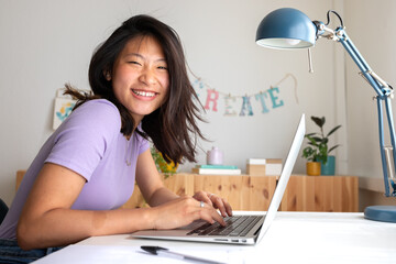 Happy, smiling teen asian female college student studying at home, doing homework using laptop looking at camera.