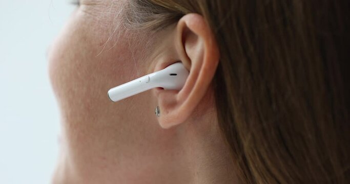 Young woman putting wireless earphone into ear close up. Gadget user lady showing using white small earbud for listening to music, video phone call. Cropped shot