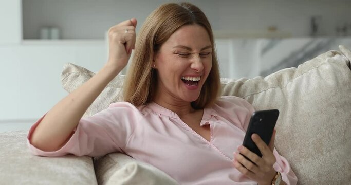 Excited happy smartphone user woman receiving good news, reading text message, throwing yes fist, making winner hand, laughing, shouting, resting on home couch, celebrating success, surprise